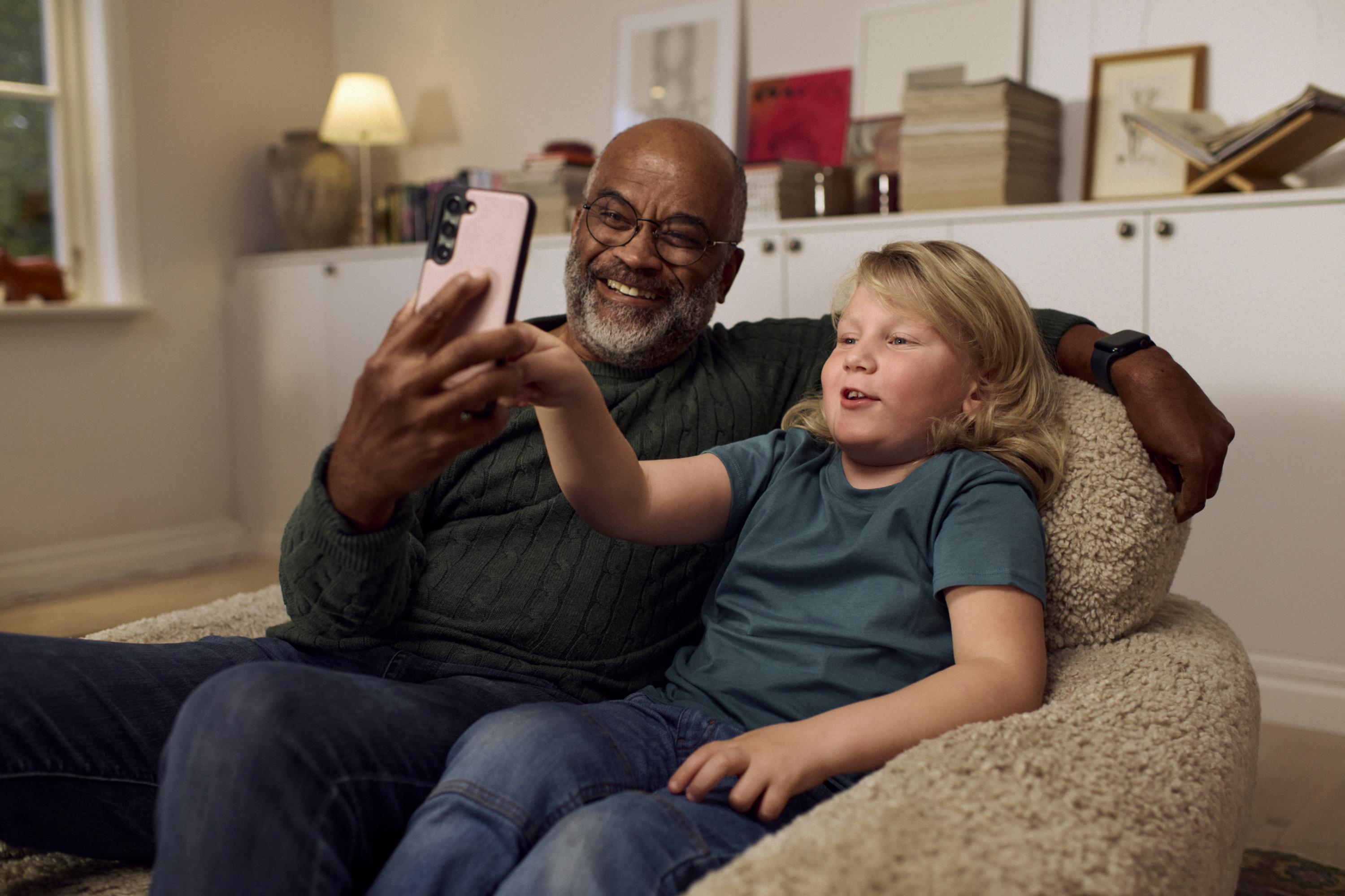 En äldre man och ett barn sitter tillsammans i en fåtölj och ler medan de tar en selfie med en mobiltelefon.
