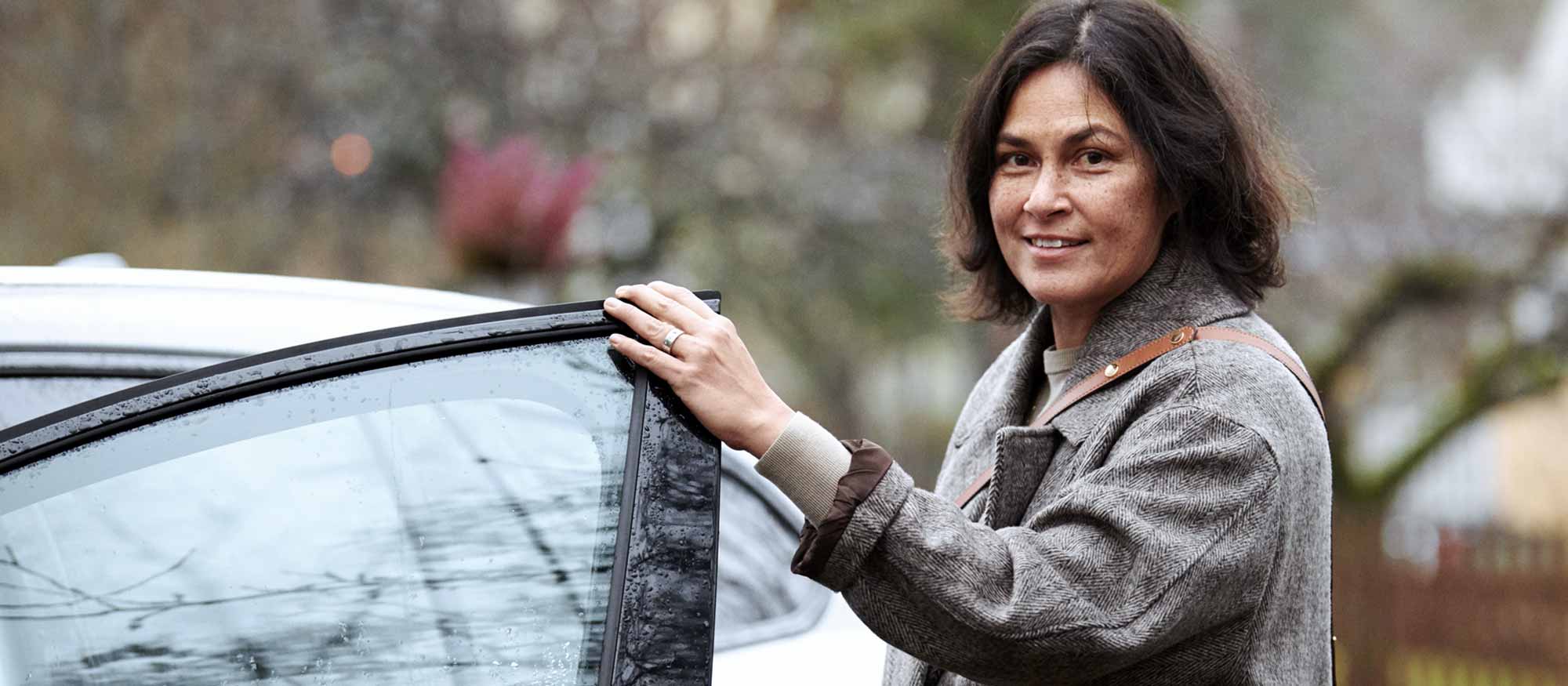 Kvinna med grå kappa håller på att kliva in i en bil en höstdag