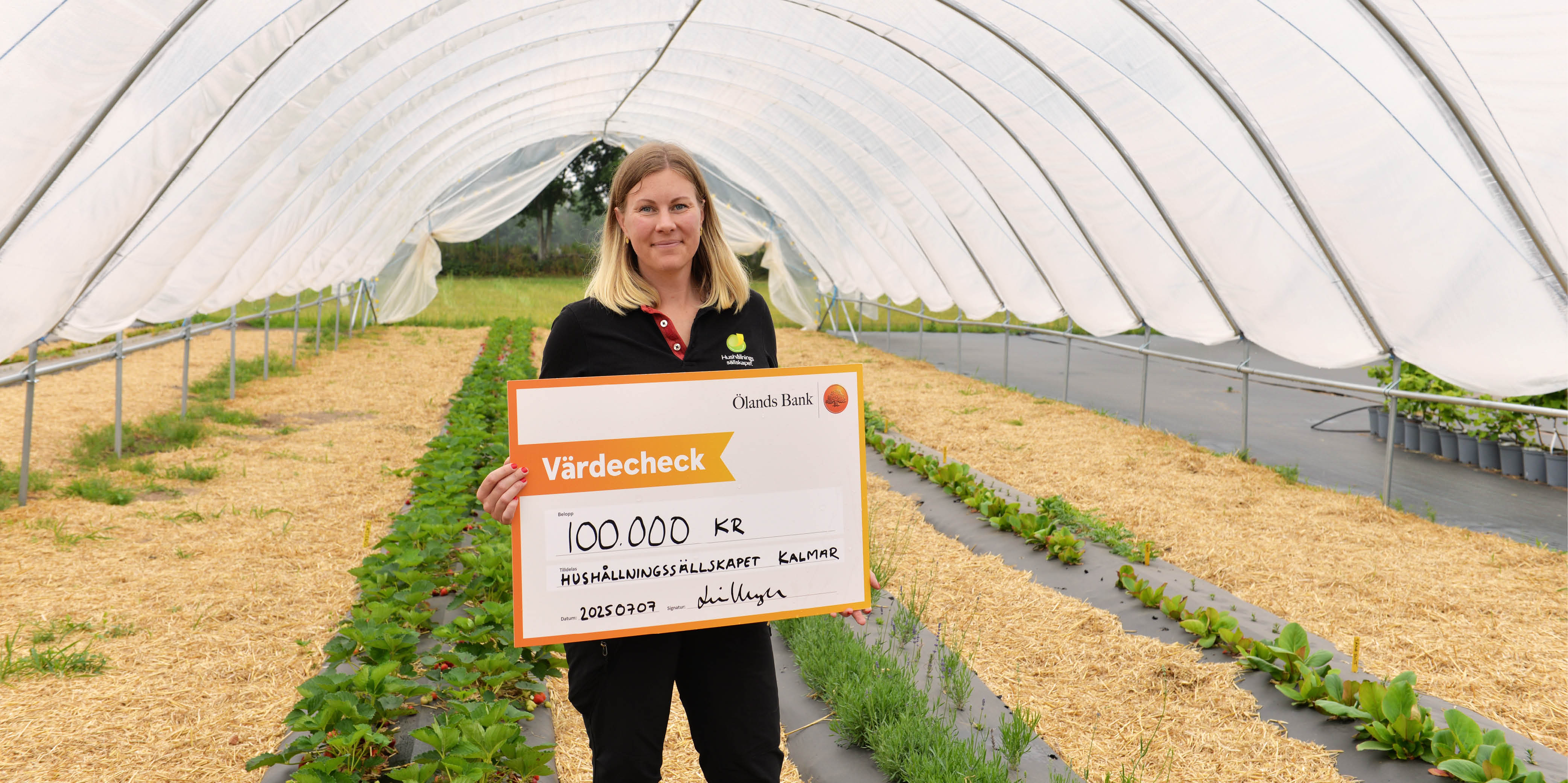 A girl stands in a large greenhouse and holding a large check with money in her hands.