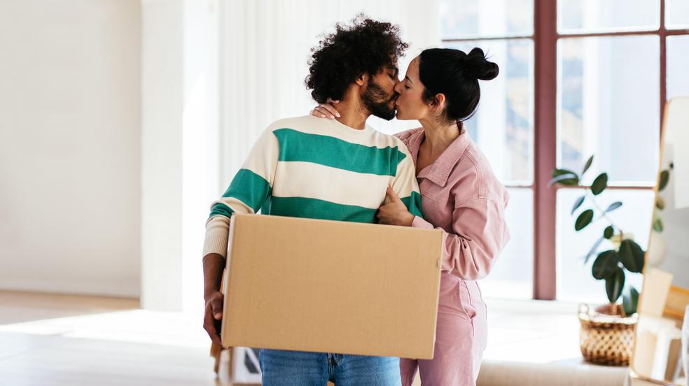 Young woman kissing black man with carton box with Kids Room inscription during relocation into new apartment